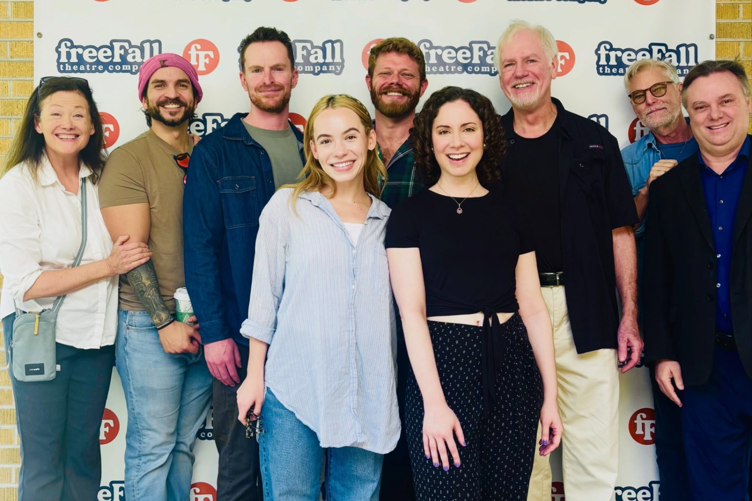 Nine members of freeFall Theatre's The Da Vinci Code company smiling in front of a freeFall Theatre Company logo backdrop