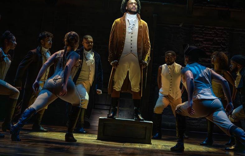 A performer in a gold velvet coat and white cravat stands on a wooden crate at center stage, surrounded by ensemble members in 18th-century costume on a dark brick set.