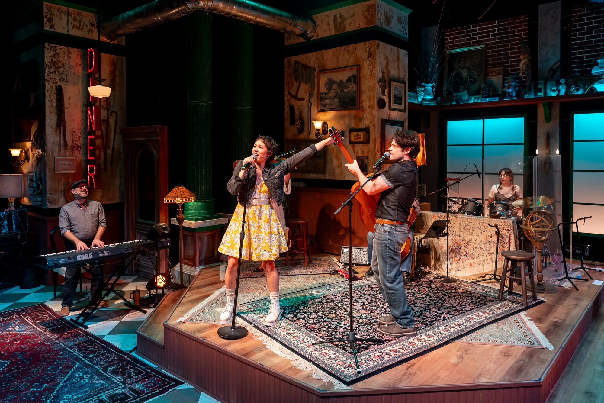 Cast members perform on a warmly lit set resembling a New York diner, with keyboards, guitar, and drums visible as the lead singers share a microphone mid-song.