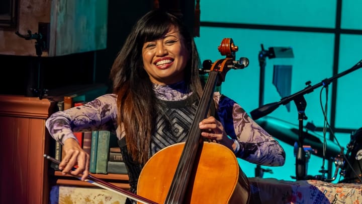Rose Mallare smiles while playing cello on stage, seated among instruments and vintage décor, with a looping pedal at her feet.