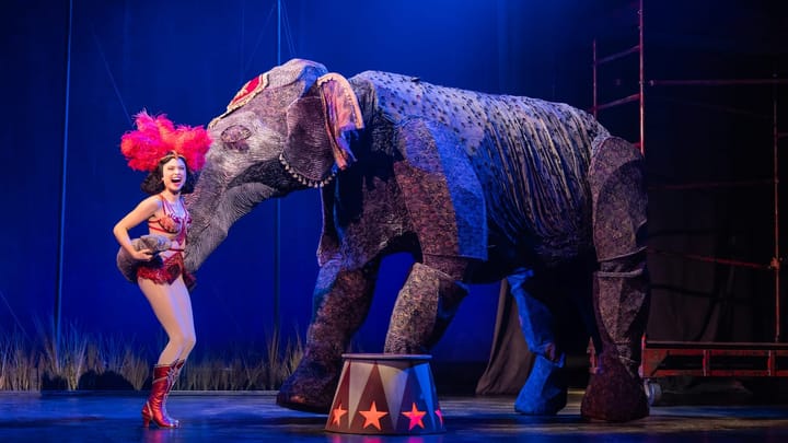 Helen Krushinski, dressed in a red circus costume with feathered headpiece, smiles beside a life-size elephant puppet during a scene from Water for Elephants.